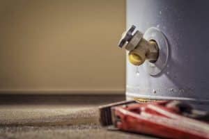A close-up of a water heater valve with a small leak, showing water dripping onto the floor. A red pipe wrench lies on the ground nearby, hinting at an upcoming water heater replacement that may require a permit for water heater work. | High 5 Plumbing, Heating & Cooling