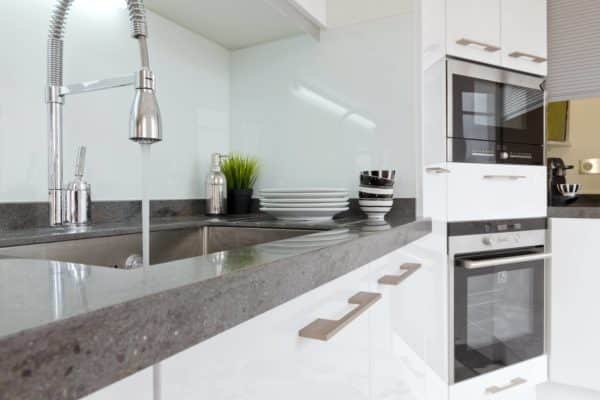 Modern kitchen with a gray countertop, stainless steel sink, and chrome pull-down faucet. Stacked white plates and bowls sit by small plants—perfect for those looking to improve water quality with stylish fixtures in white cabinetry. | High 5 Plumbing, Heating & Cooling