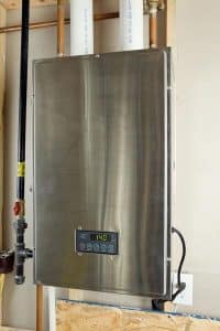 A stainless steel tankless water heater, an energy-efficient plumbing appliance, is wall-mounted with visible water pipes at the top and a digital control panel on the front displaying a 140°F temperature reading. | High 5 Plumbing, Heating & Cooling
