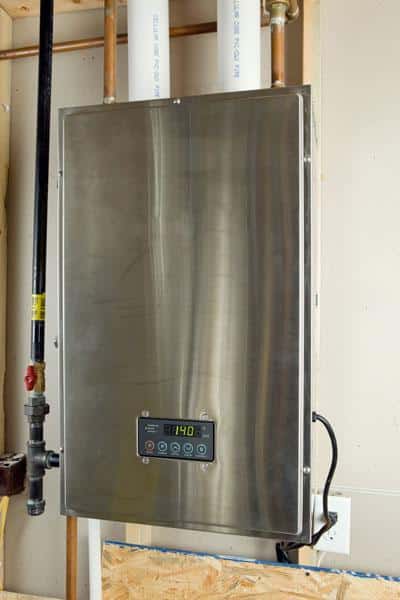 A stainless steel tankless water heater, an energy-efficient plumbing appliance, is wall-mounted with visible water pipes at the top and a digital control panel on the front displaying a 140°F temperature reading. | High 5 Plumbing, Heating & Cooling