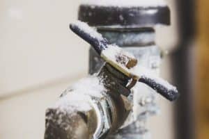 A close-up of an outdoor water faucet covered in snow and ice, highlighting cold winter weather and the risk of frozen pipes. The metallic faucet with a blue handle serves as a reminder of what to do when pipes freeze. | High 5 Plumbing, Heating & Cooling