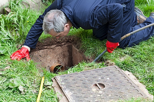 A man wearing red gloves and a blue jumpsuit kneels on grass in Denver, inspecting an open rectangular hole with a pipe inside, possibly performing a drain repair. A metal cover lies nearby on the ground. | High 5 Plumbing, Heating & Cooling
