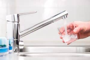 A hand is holding a glass under a modern kitchen faucet, filling it with running water—an ideal moment to consider water purifier installation in this clean, bright kitchen with a stainless steel sink. | High 5 Plumbing, Heating & Cooling