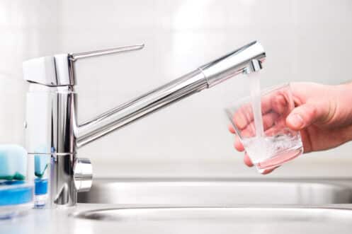 A hand is holding a glass under a modern kitchen faucet, filling it with running water—an ideal moment to consider water purifier installation in this clean, bright kitchen with a stainless steel sink. | High 5 Plumbing, Heating & Cooling