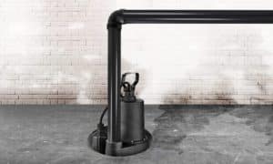 A black sump pump with high sump pump capacity is installed in a circular pit on a concrete floor, connected to black PVC pipes, with a white brick wall in the background. | High 5 Plumbing, Heating & Cooling