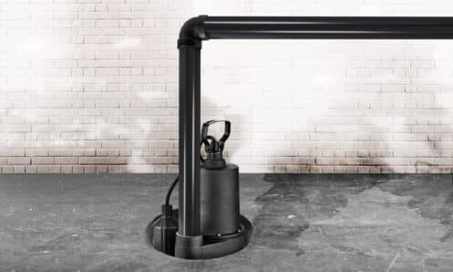 A black sump pump with high sump pump capacity is installed in a circular pit on a concrete floor, connected to black PVC pipes, with a white brick wall in the background. | High 5 Plumbing, Heating & Cooling