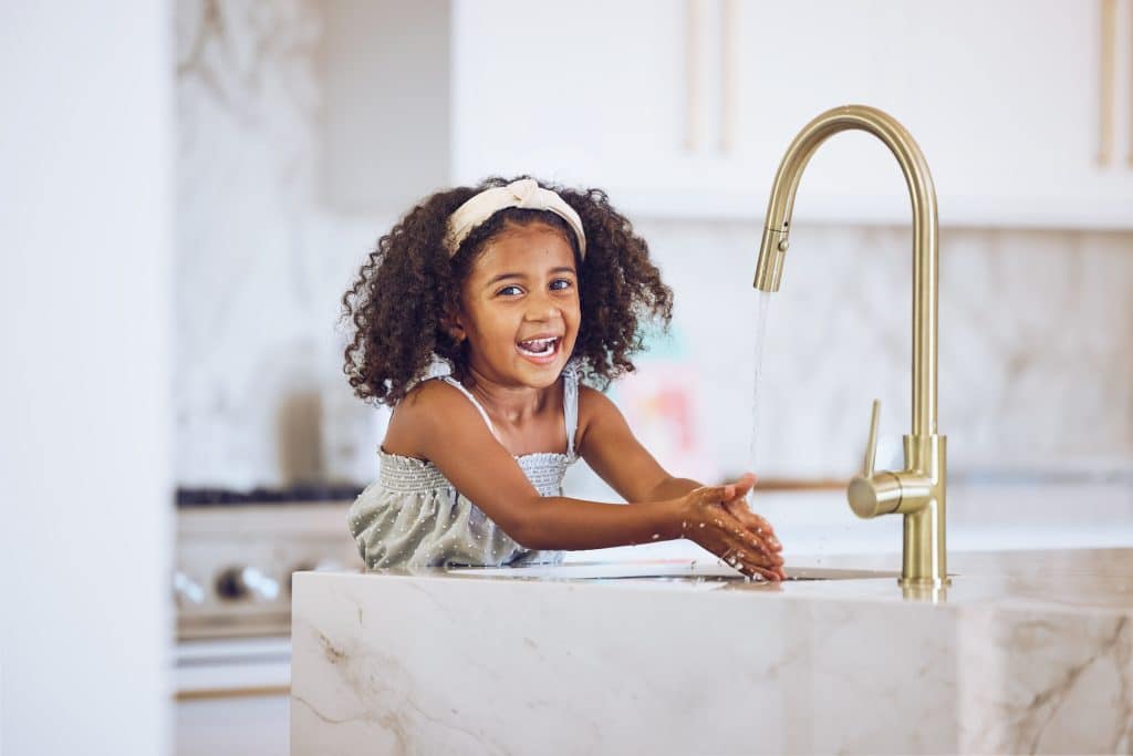 A young girl with curly hair and a headband is smiling while washing her hands under a running faucet with water filtration at a marble kitchen sink. The background shows a modern kitchen with white cabinets. | High 5 Plumbing, Heating & Cooling