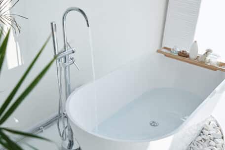 A modern white bathtub is being filled with water from a chrome faucet. Reflecting bathroom trends 2024, a wooden tray with bath items sits on the tub's edge, with white decorative stones nearby and a green plant in the foreground. | High 5 Plumbing, Heating & Cooling