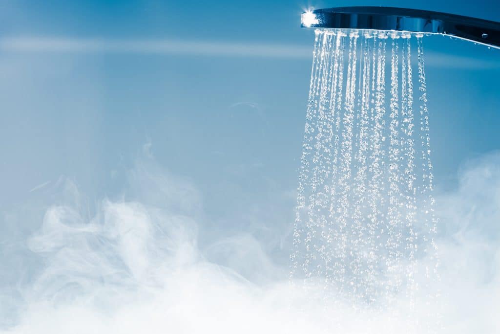 A close-up of a showerhead spraying water, with visible steam rising below, indicating hot water in use. The soft blue background adds to the clean, fresh atmosphere—perfect inspiration for luxurious bathroom or shower upgrades. | High 5 Plumbing, Heating & Cooling
