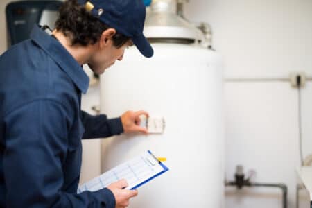 A person in a blue uniform and cap inspects a white tankless water heater, holding a clipboard and adjusting a control on the unit in a utility room while troubleshooting a beeping alert. | High 5 Plumbing, Heating & Cooling