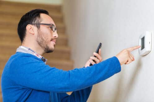 A man wearing glasses and a blue sweater is holding a smartphone and adjusting a wall-mounted thermostat, possibly checking for AC repair needs. There are stairs in the background. | High 5 Plumbing, Heating & Cooling
