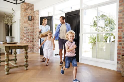 A family of four—two adults and two young children—enter a bright, modern home with wooden floors and large windows, designed to improve HVAC efficiency. The cheerful children run ahead, while the adults walk behind them, all appearing happy. | High 5 Plumbing, Heating & Cooling