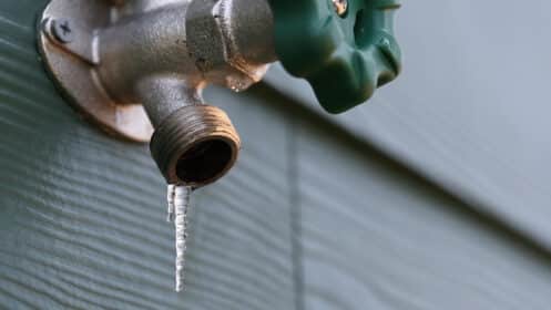 A close-up of an outdoor metal water faucet with a green handle, attached to a wall, with a small icicle hanging from the spout—an early sign of winter and a reminder of the risk of frozen pipes. | High 5 Plumbing, Heating & Cooling