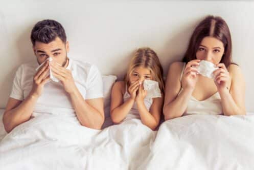 A man, a young girl, and a woman sit together in bed, holding tissues to their noses as if sneezing, likely due to dry winter air in Denver. All three are under white blankets and wearing white tops. | High 5 Plumbing, Heating & Cooling
