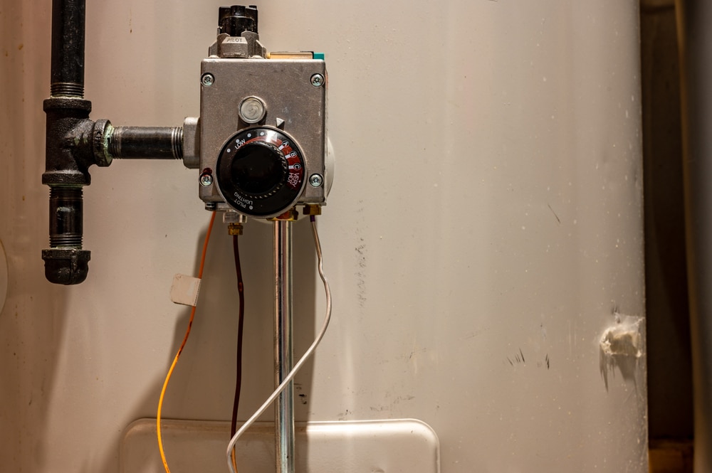 A close-up view of a residential water heater thermostat with wires attached, mounted on a white water heater tank. Black piping and the temperature dial for adjusting hot water heater settings are also visible. | High 5 Plumbing, Heating & Cooling