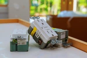 Three electrical outlets, including a GFCI outlet for Load vs Line Wiring difference, rest on a white surface in a room under construction with blurred green scenery visible through windows in the background. | High 5 Plumbing, Heating & Cooling