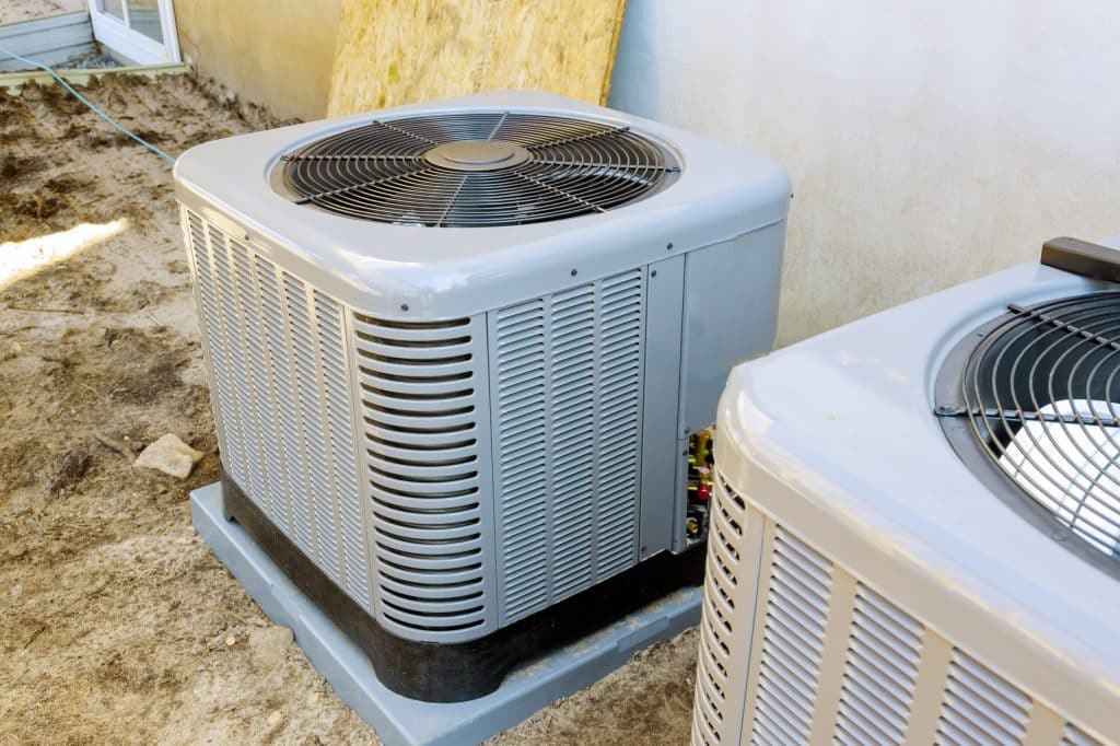 Two outdoor air conditioning units are installed on concrete pads next to a building. Upgrading to units with higher air filter efficiency or improved MERV filter ratings can further enhance indoor air quality. The ground is bare soil, and a window is partially visible. | High 5 Plumbing, Heating & Cooling
