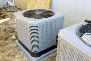 Two outdoor air conditioning units are installed on concrete pads next to a building. Upgrading to units with higher air filter efficiency or improved MERV filter ratings can further enhance indoor air quality. The ground is bare soil, and a window is partially visible. | High 5 Plumbing, Heating & Cooling