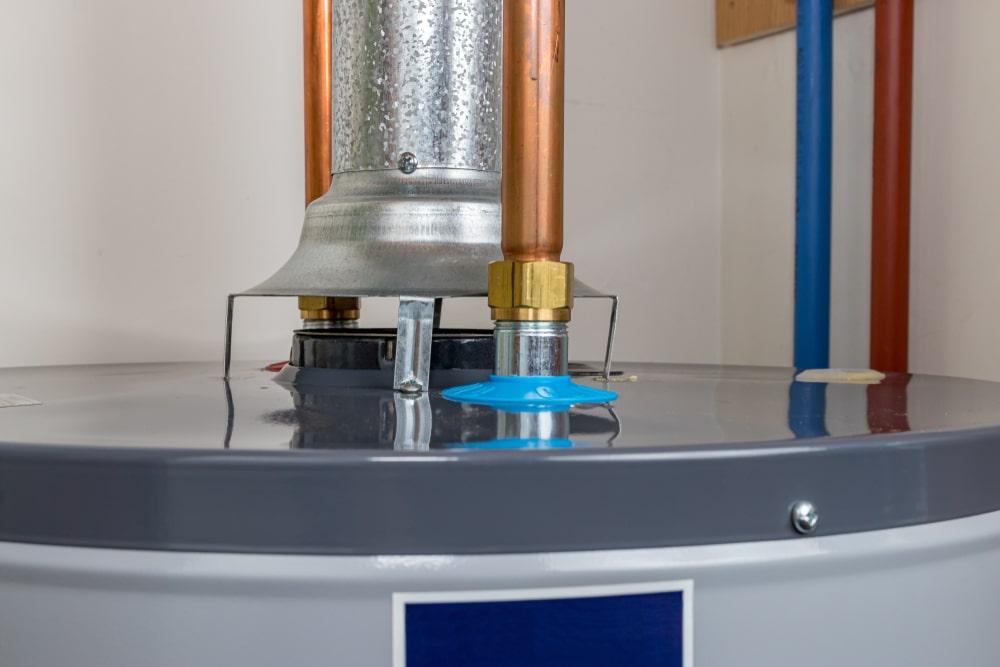A close-up view of the top of a gas water heater shows metal and copper pipes, a vent, and a blue plastic cap, with a white wall in the background. | High 5 Plumbing, Heating & Cooling