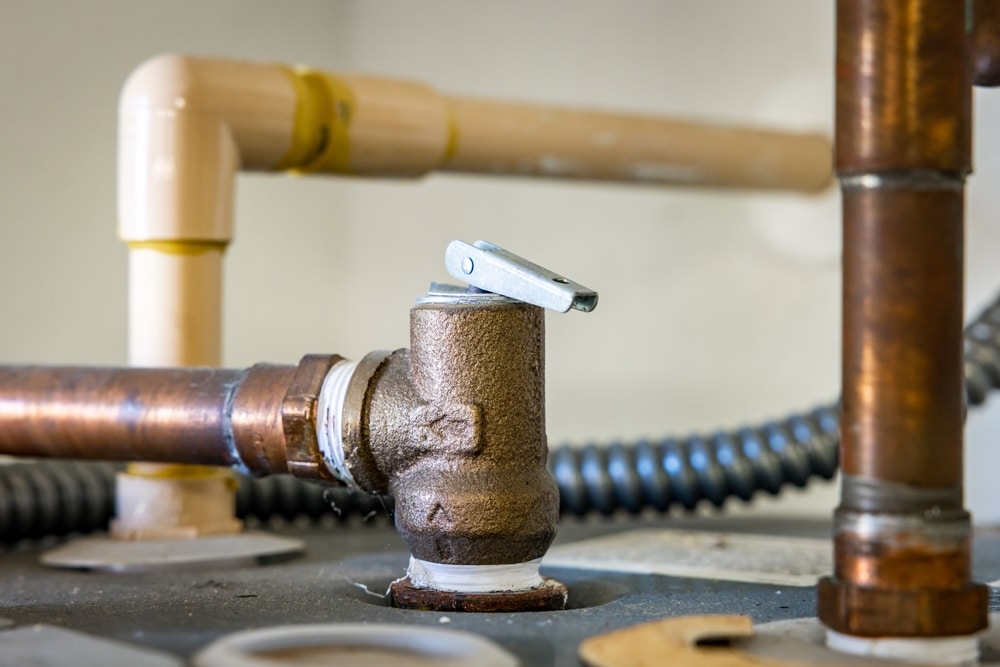 Close-up of a water heater pressure relief valve attached to a copper pipe, with additional plastic and metal pipes in the background. The valve is installed on a flat metal surface. | High 5 Plumbing, Heating & Cooling