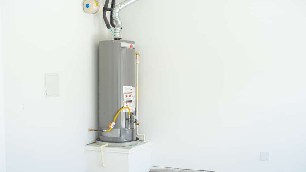 A gray water heater is installed in the corner of a white room, elevated on a small platform, with pipes and ventilation attached. The unfinished room highlights the appliance, raising questions about water heater longevity in such spaces. | High 5 Plumbing, Heating & Cooling
